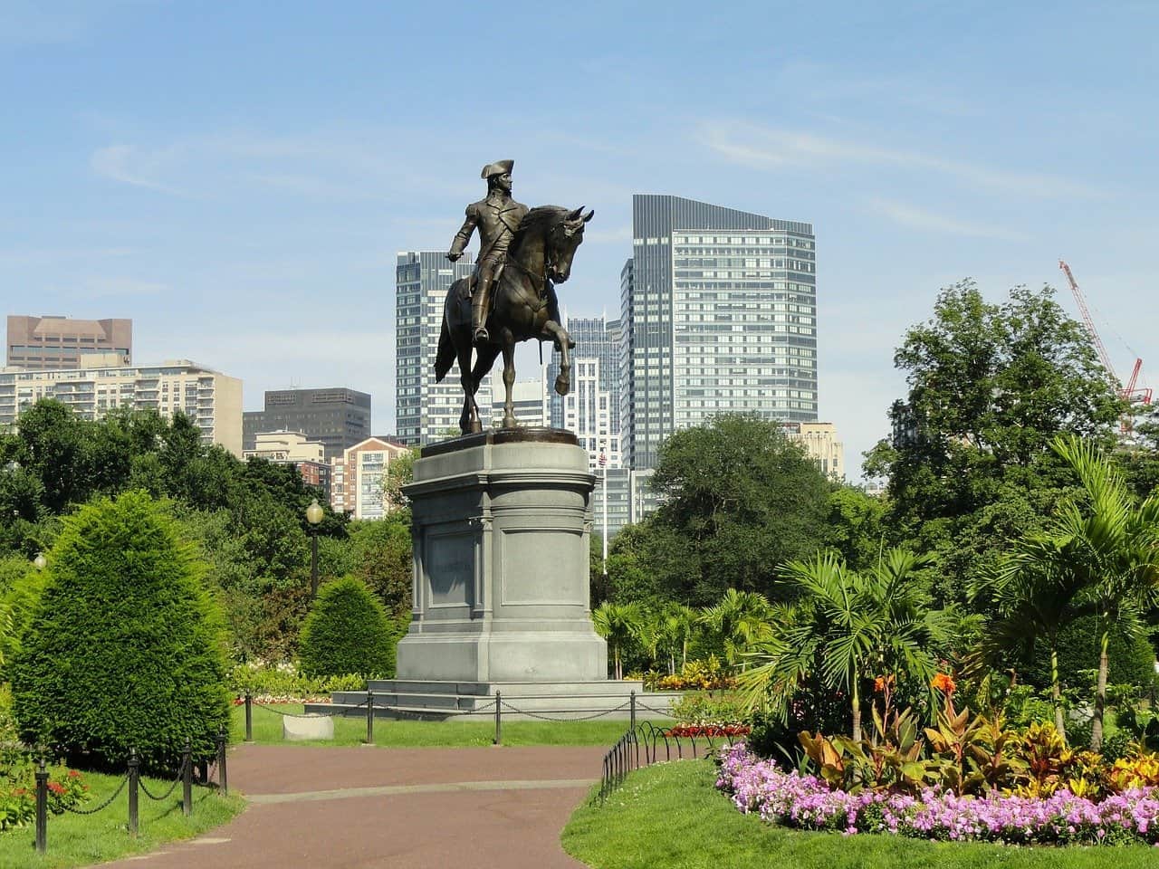 Paul Revere statue in Boston Commons, with Boston buildings in the background and green trees surrounding the statue.