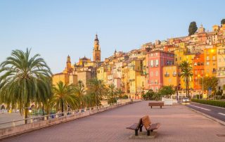 Promenade along the water with colorful buildings and palm trees in Menton, France.